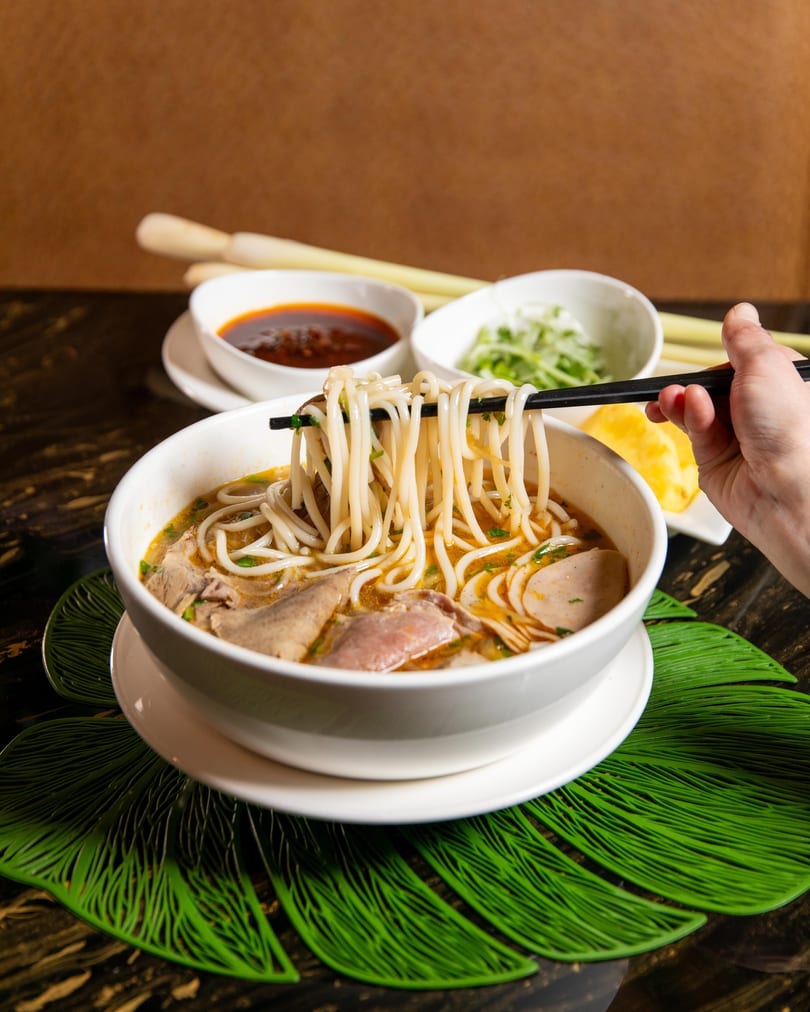 A hand uses chopsticks to lift noodles from a bowl of Vietnamese bun bo hue soup.
